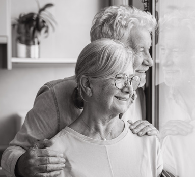 A black and white photo of an elderly couple looking out a window, smiling and embracing